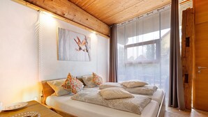 Une chambre avec un plafond en bois, un lit recouvert d’une literie à motifs, une table de chevet sur laquelle se trouve une assiette de fruits, et un tableau accroché au mur.
