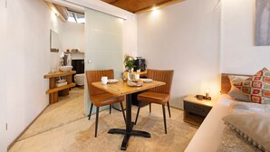 Une pièce de taille modeste, avec une table à manger en bois, deux chaises, une petite table de chevet avec une lampe, un canapé agrémenté de coussins à motifs et un plafond en bois.
