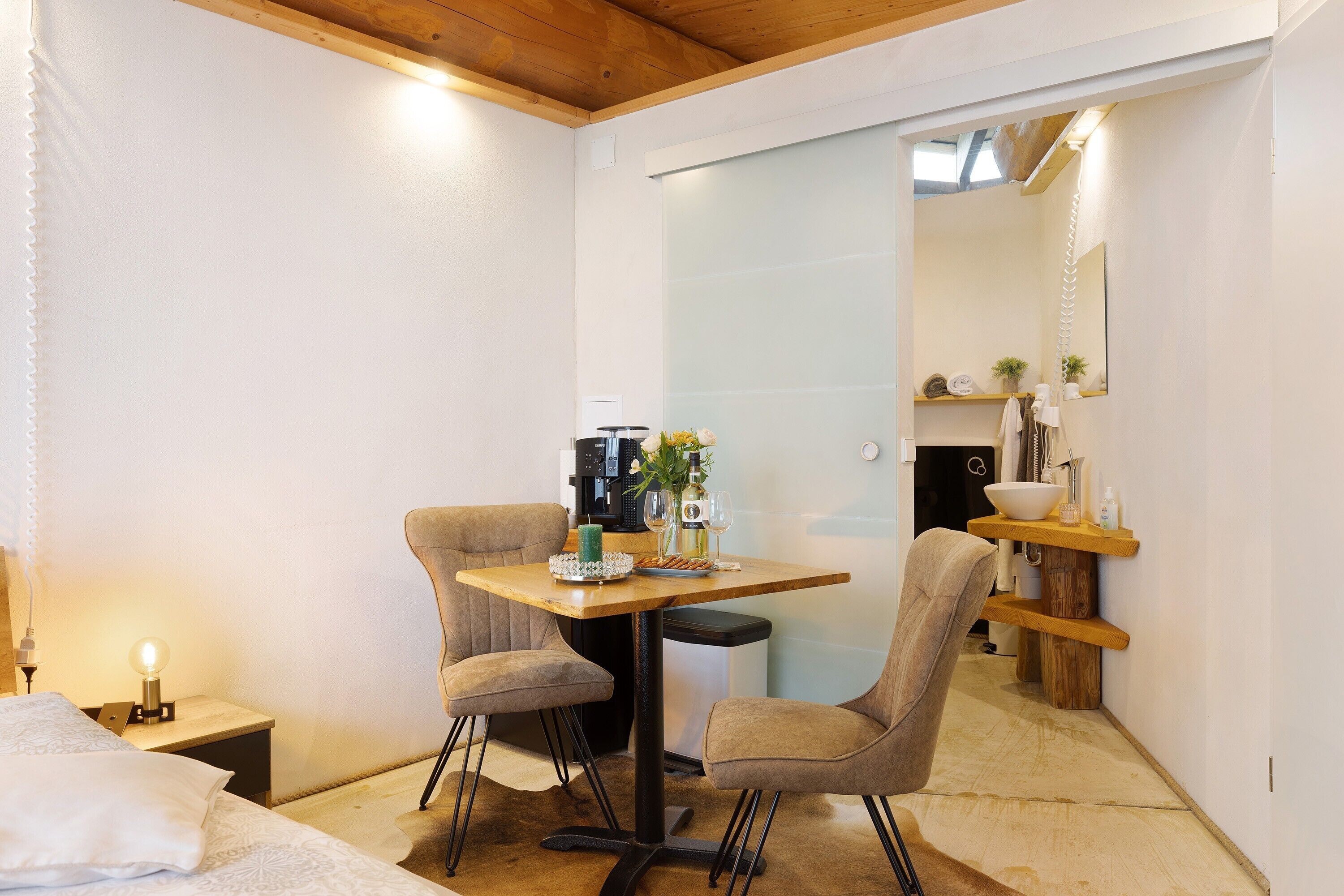 Een knus, klein kamertje met een bed, een tafel met stoelen, een koffieapparaat en een spiegel.