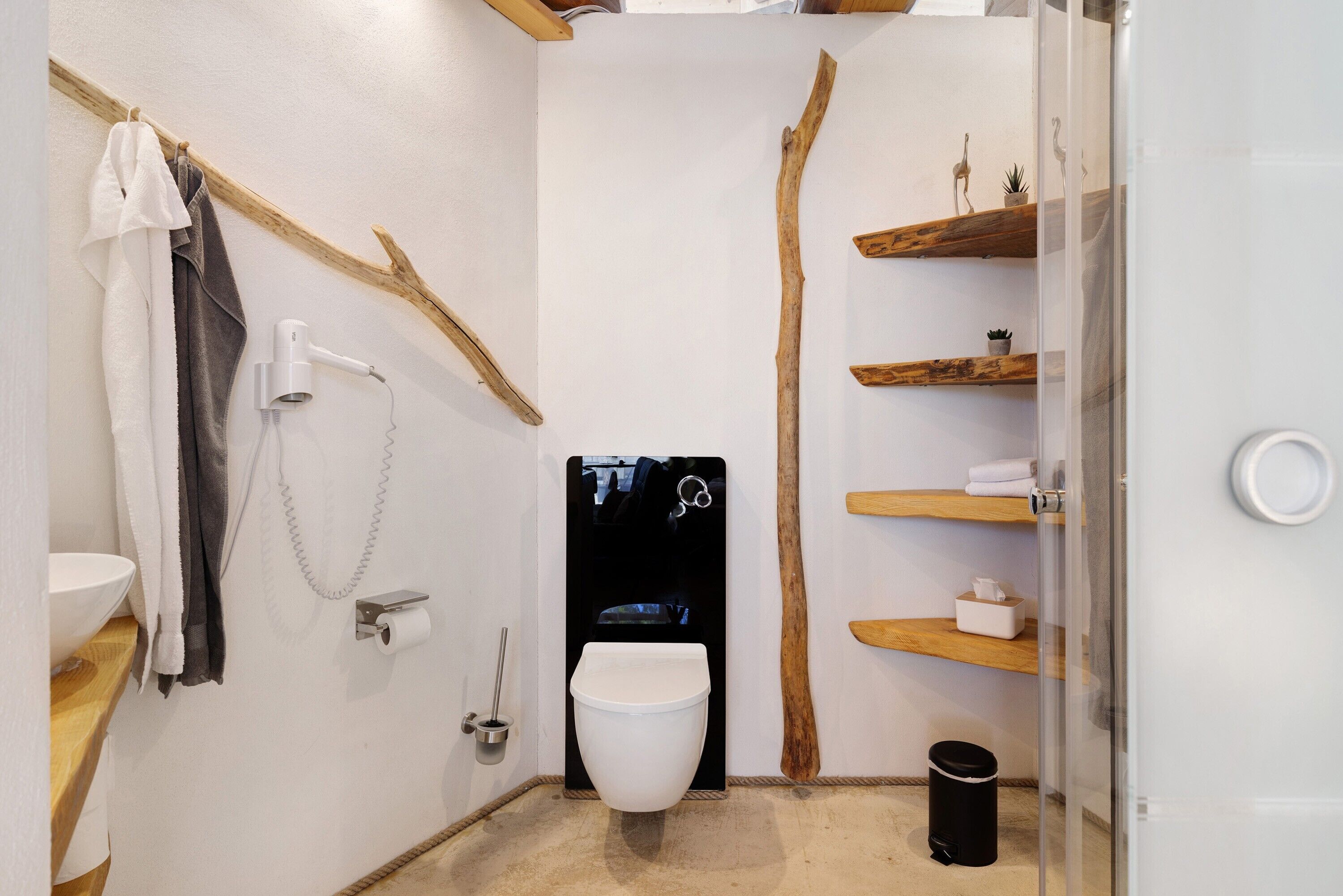 Une salle de bain équipée d’un WC, d’un porte-serviettes, d’un sèche-cheveux, d’une étagère en bois et d’une poubelle.