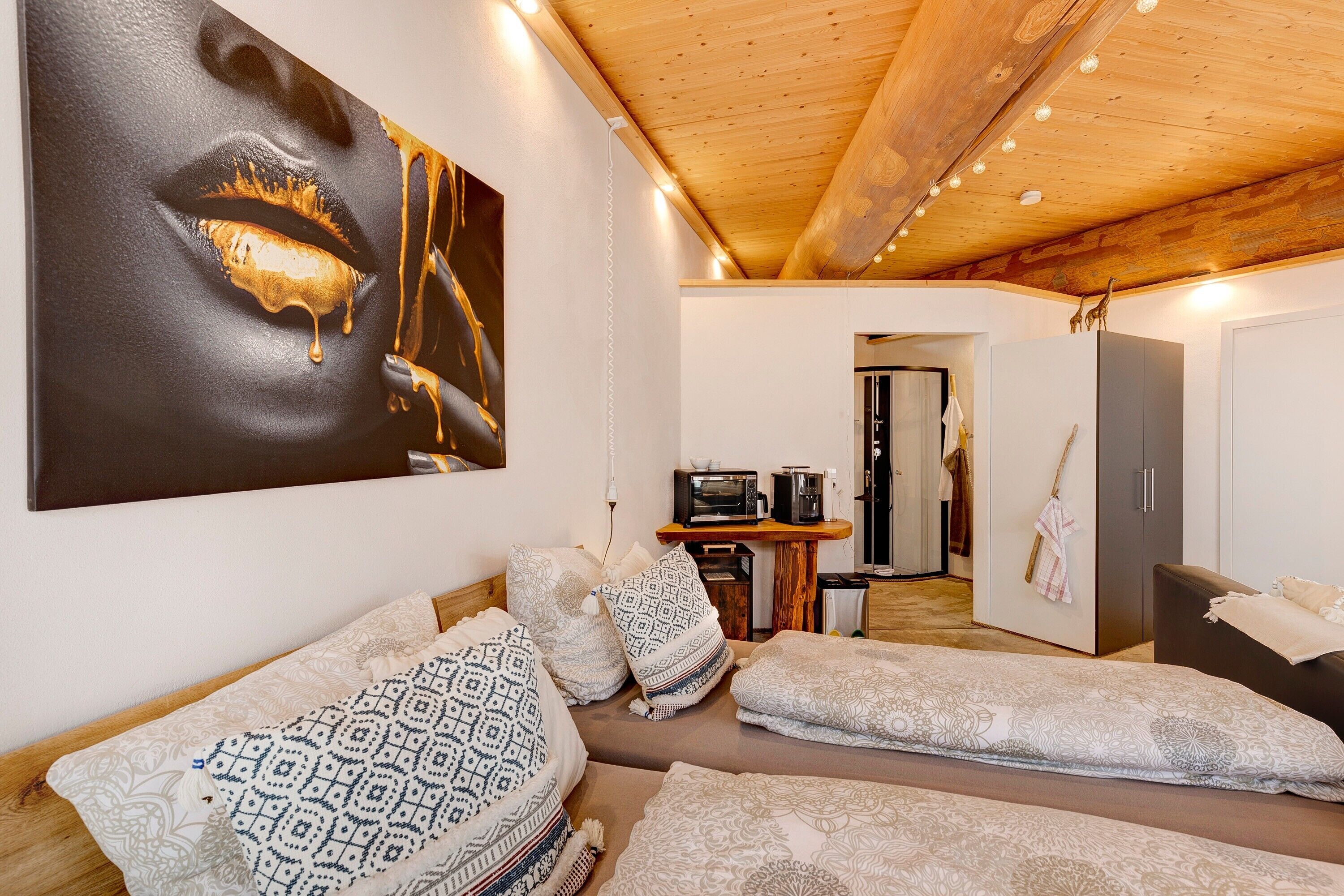 Een modern interieur met een houten plafond, een groot abstract schilderij, een bank met gekleurde kussens, een eethoek met een magnetron en een kledingkast.