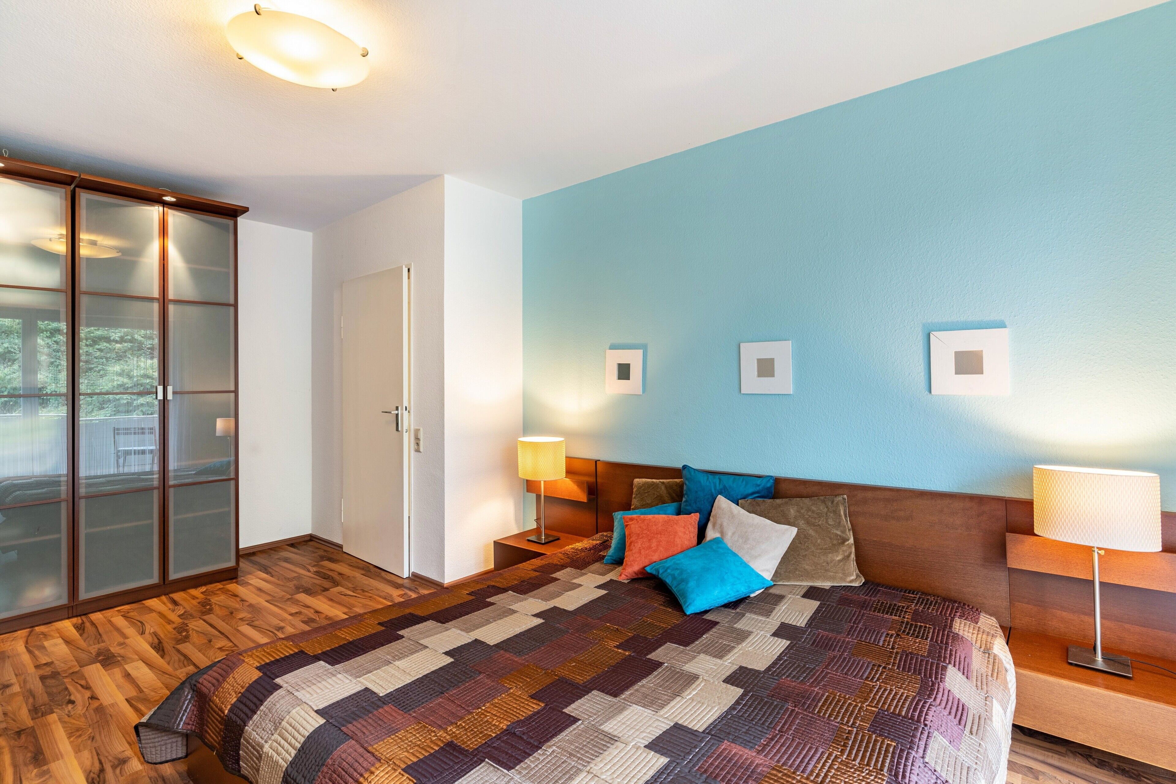 Quarto com uma cama grande, um guarda-roupa com portas de vidro, uma mesinha de cabeceira com abajur e um ponto de luz fixado na parede.