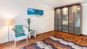 A room with a wooden floor, a white chair with a blue cushion, a small white side table with a vase, a painting of a beach scene, and a large wooden cabinet with glass doors. - "Am Paradies" Apartment – Relaxation in Baden-Baden (Baden-Baden)