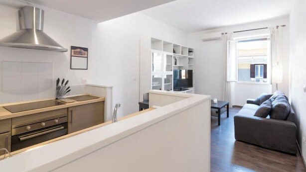 Eine moderne Küche mit Einbauofen, einer Edelstahl-Dunstabzugshaube und einer weißen Arbeitsplatte. Es gibt eine Essecke mit einem grauen Sofa und einem kleinen Tisch. Der Raum hat weiße Wände und ein großes Fenster, das für natürliches Licht sorgt.