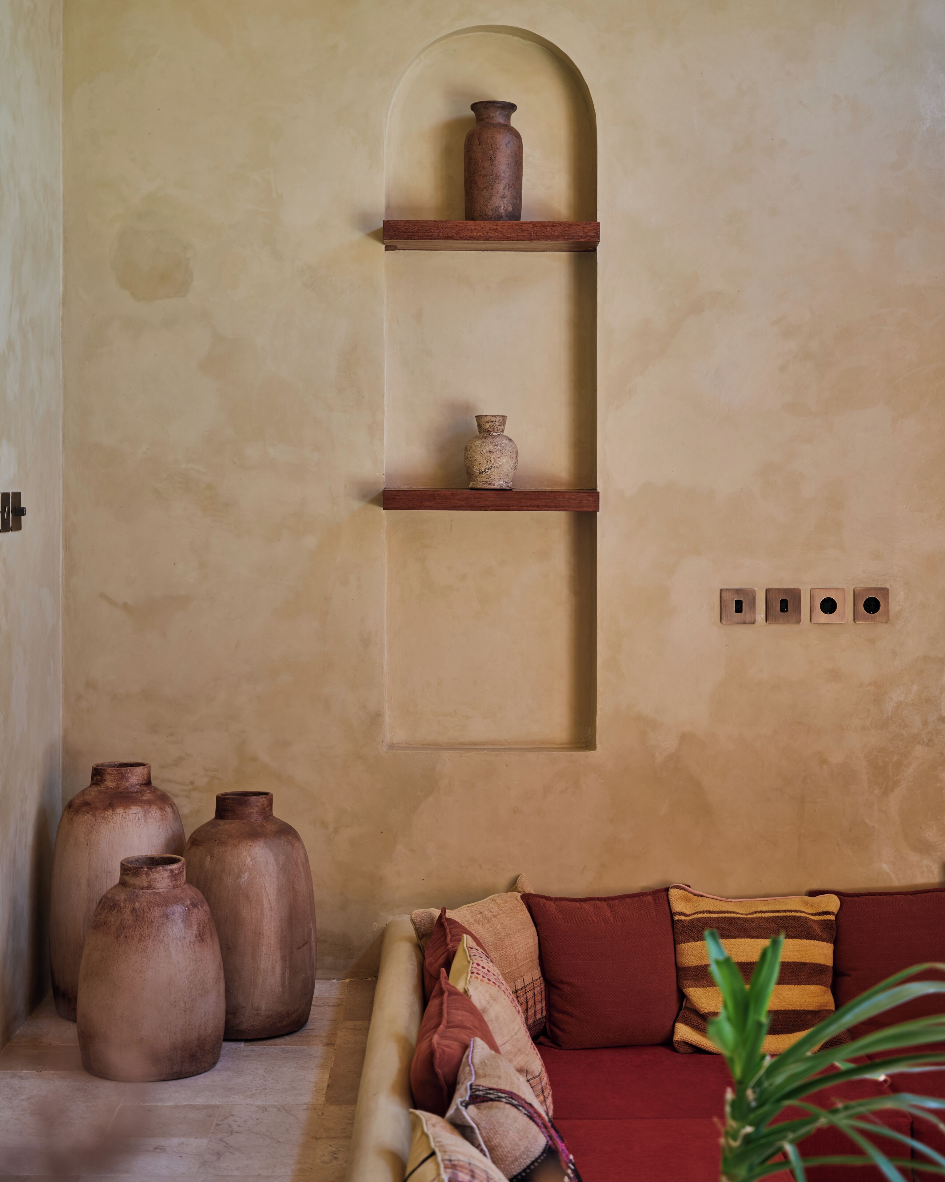 A room with a beige textured wall, a wooden shelf with three pottery pieces, a red sofa with patterned cushions, and a plant in the foreground.