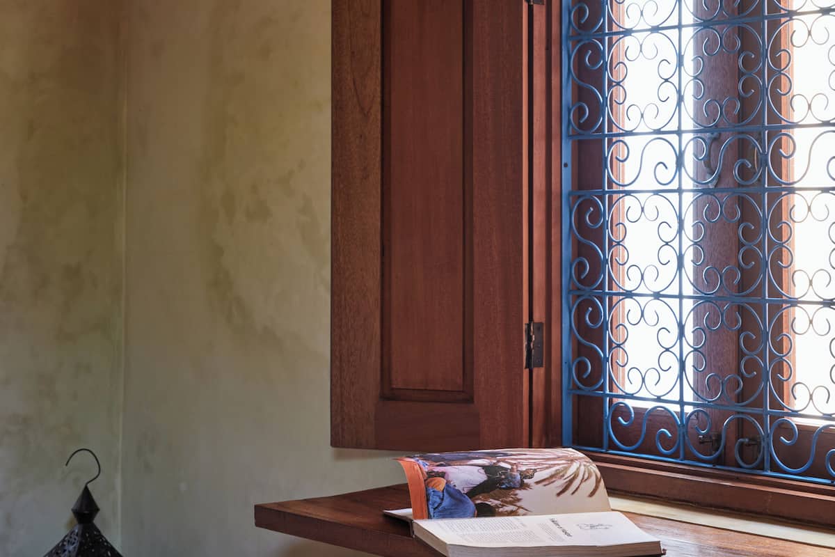 Ein Zimmer mit einem Holztisch, einem Fenster mit verzierten Metallgitterstäben, einem braunen Ledersitzhocker und einem an der Wand befestigten Regal mit Büchern und einer Lampe.