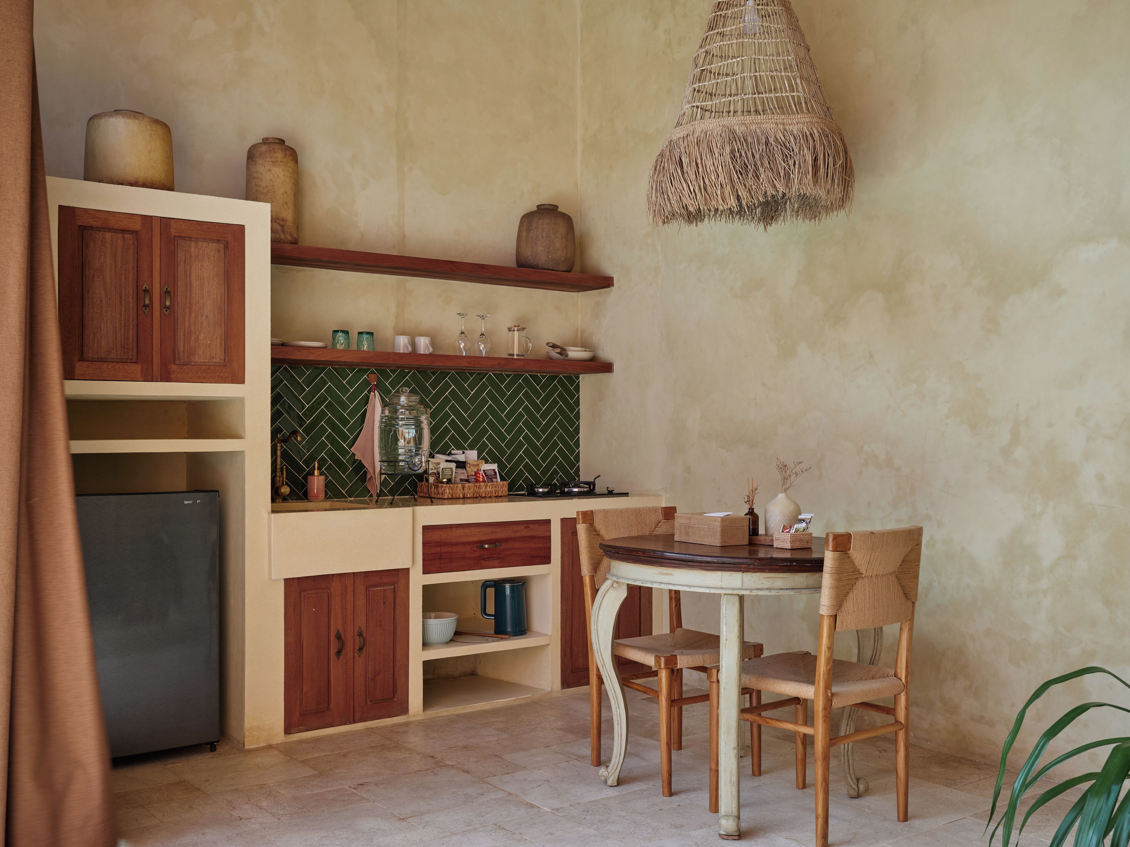 Une cuisine compacte avec des meubles en bois, un réfrigérateur encastré, une table de salle à manger ronde avec des chaises en osier et un luminaire suspendu.