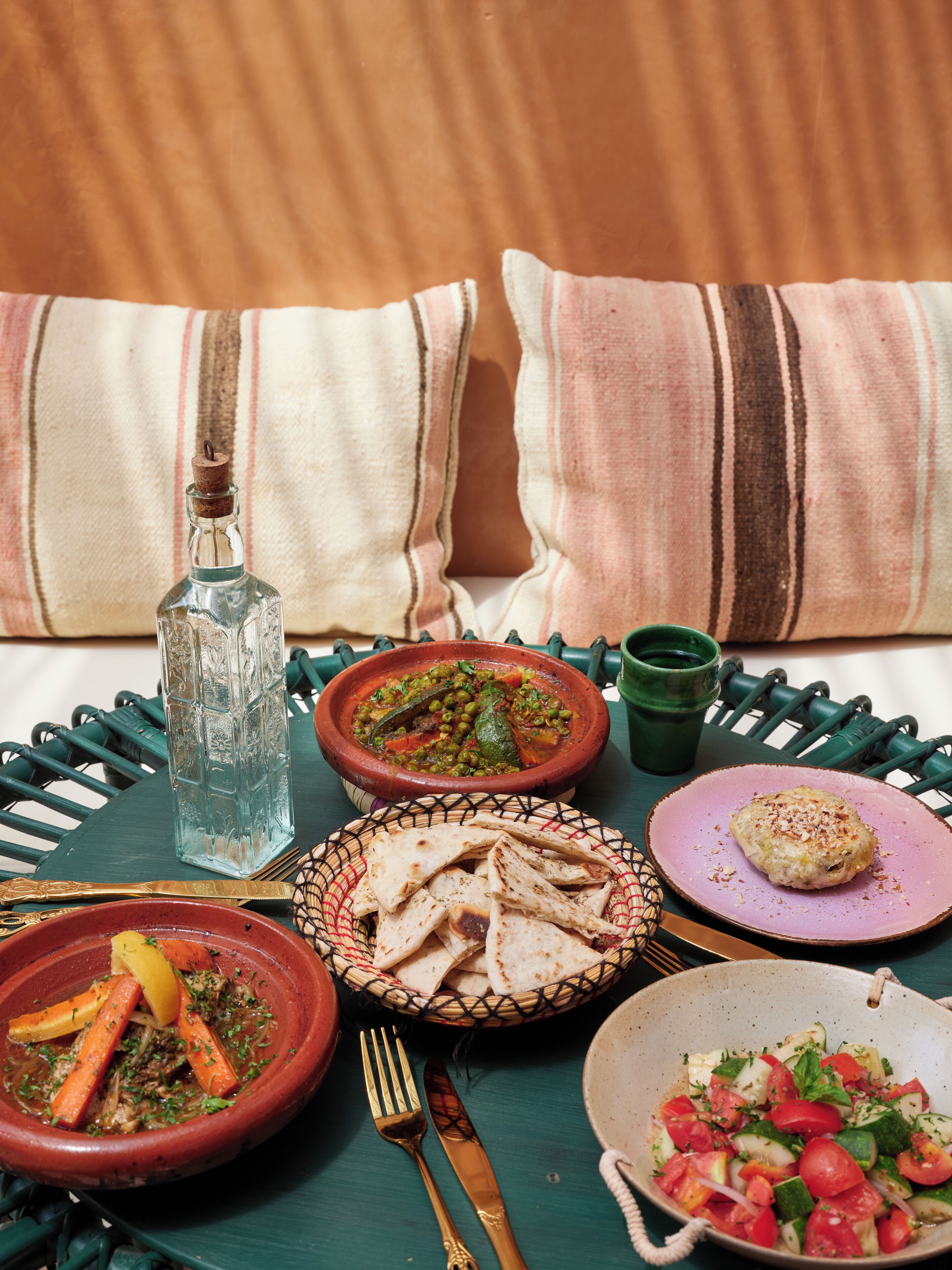 A dining table with various dishes, including a bowl of stew, a plate of flatbread, and a salad. There are also a bottle and a cup on the table.