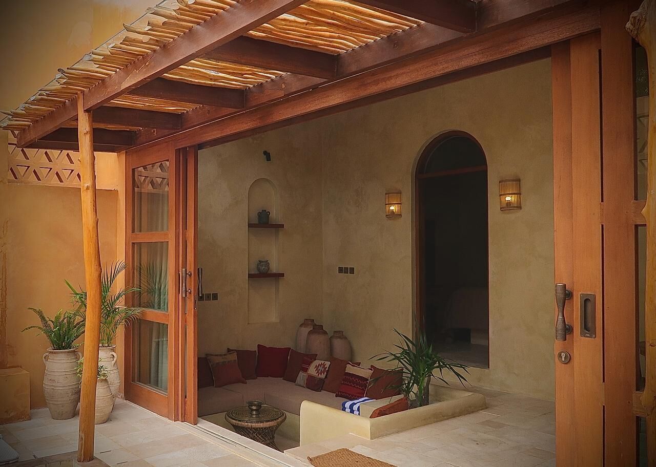 A cozy interior with a wooden pergola, a seating area with cushions, and a large arched doorway.