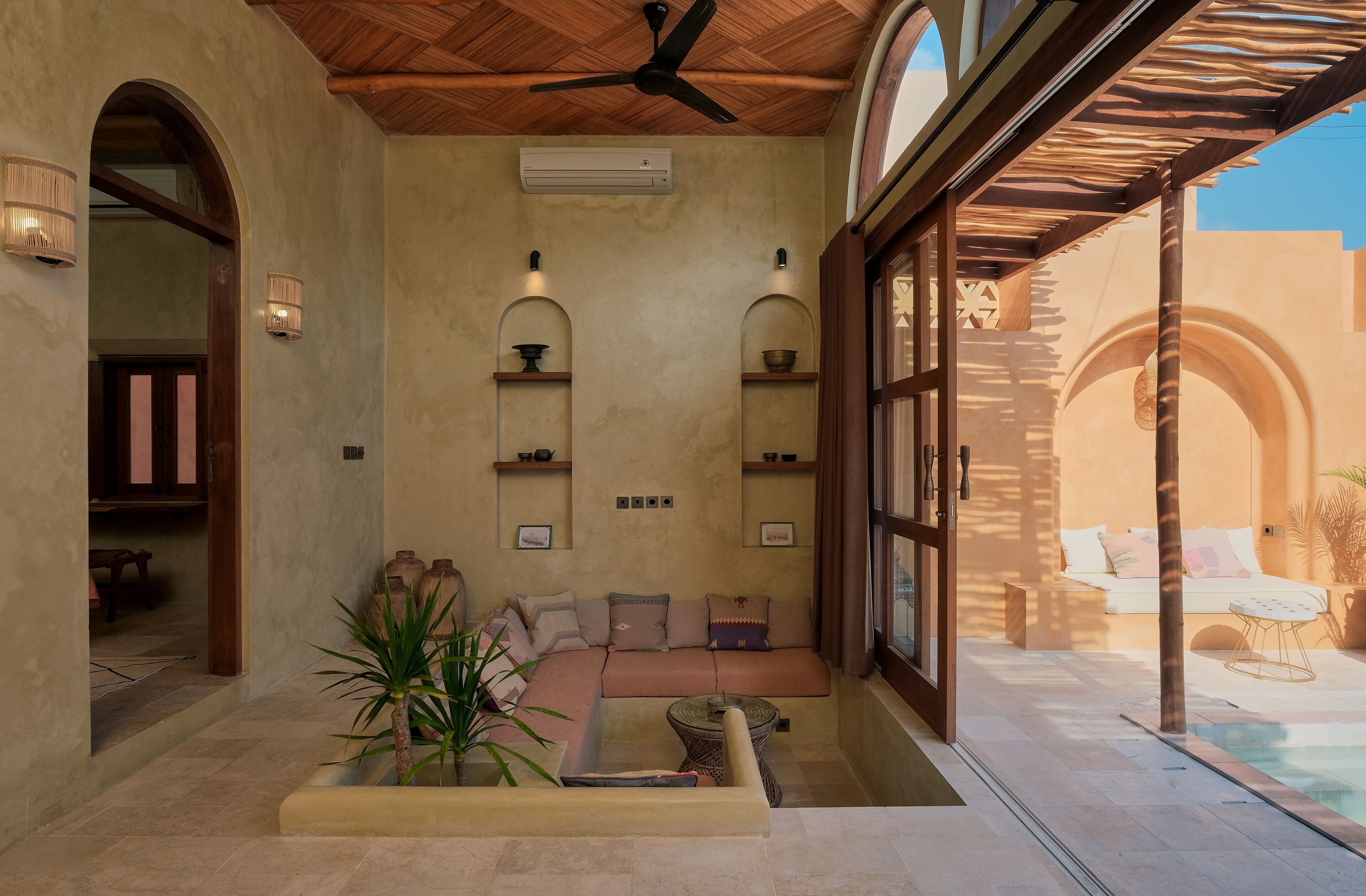 Un rincón interior acogedor con un sofá, una mesita y una planta en maceta. Se observan estanterías con objetos decorativos y un ventilador de techo.