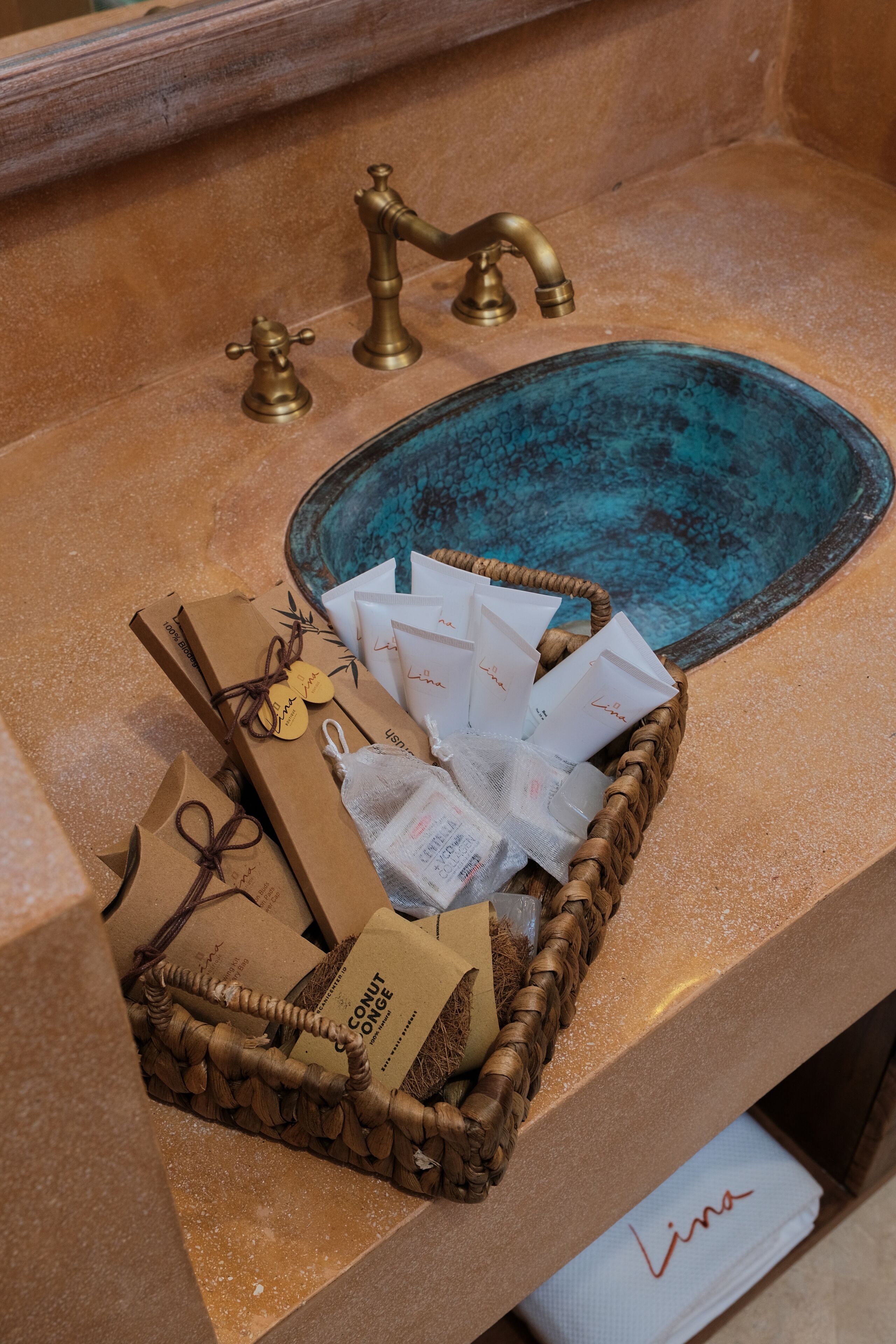 A bathroom sink with a blue basin, a brass faucet, and a wicker basket containing toiletries and a towel labeled "Lina".