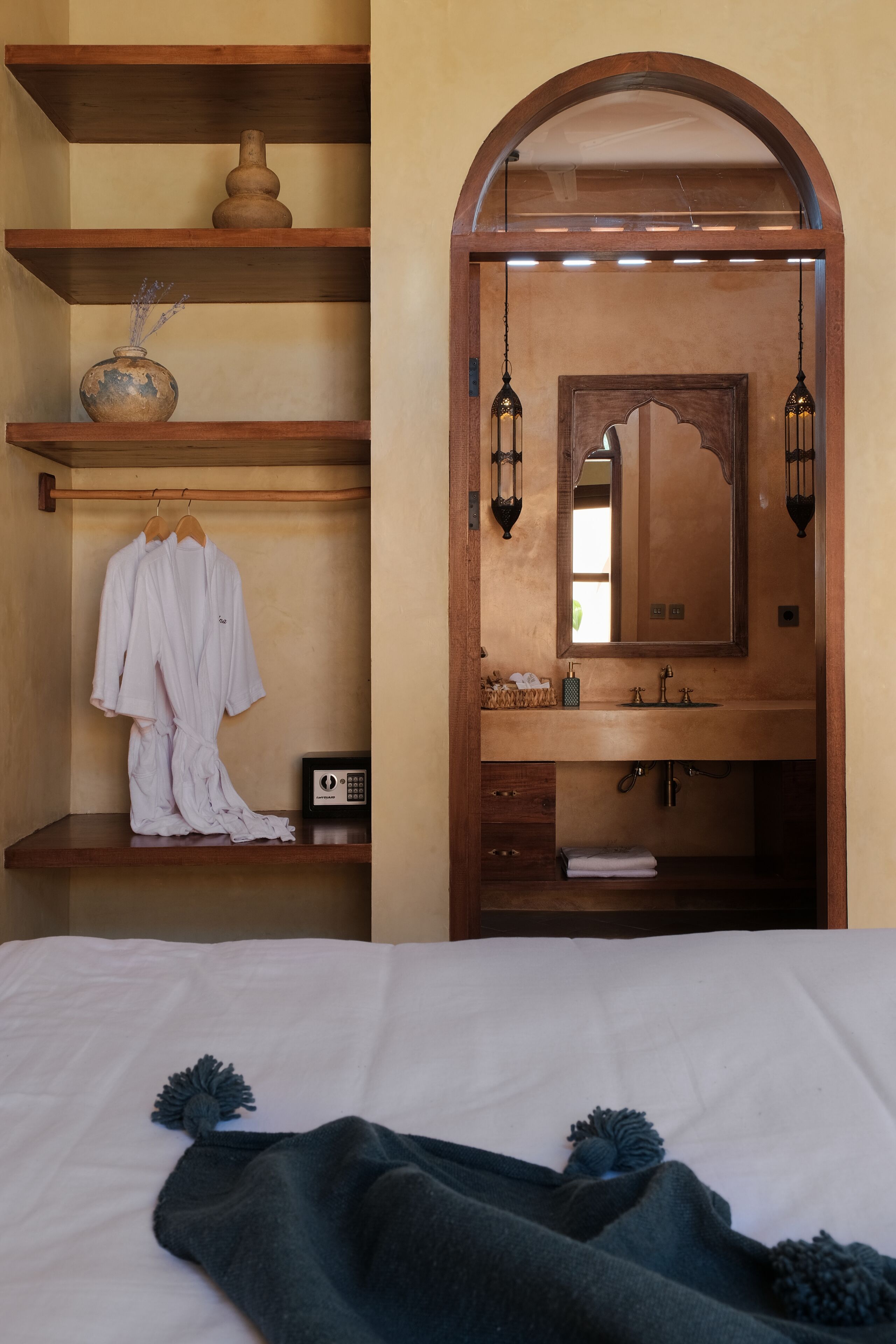 A bedroom with a wooden vanity, a mirror, and a hanging lamp. There is a bed with a white comforter and a dark blanket.