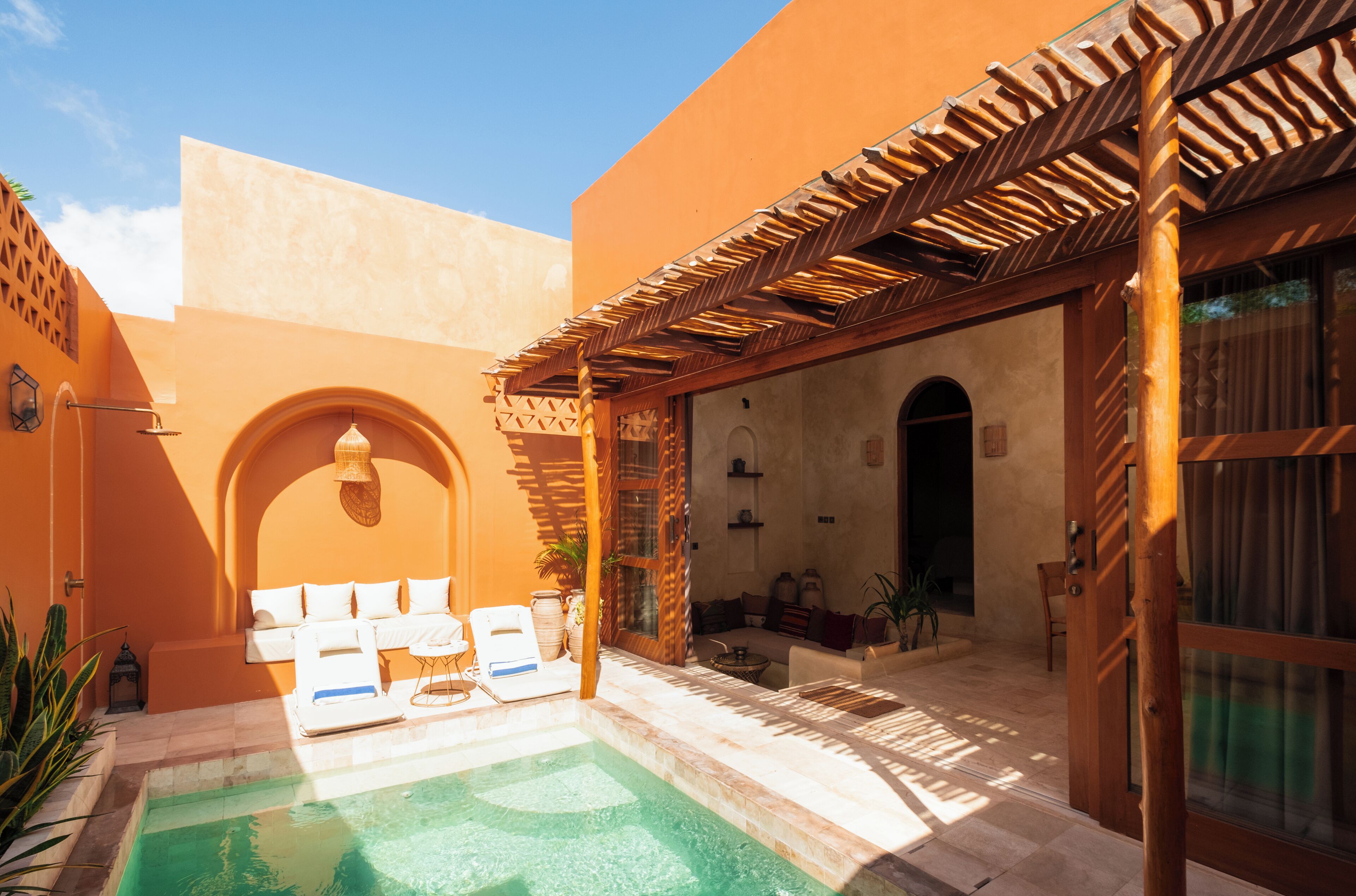 A courtyard with a pool, a wooden pergola, and a seating area with cushions.