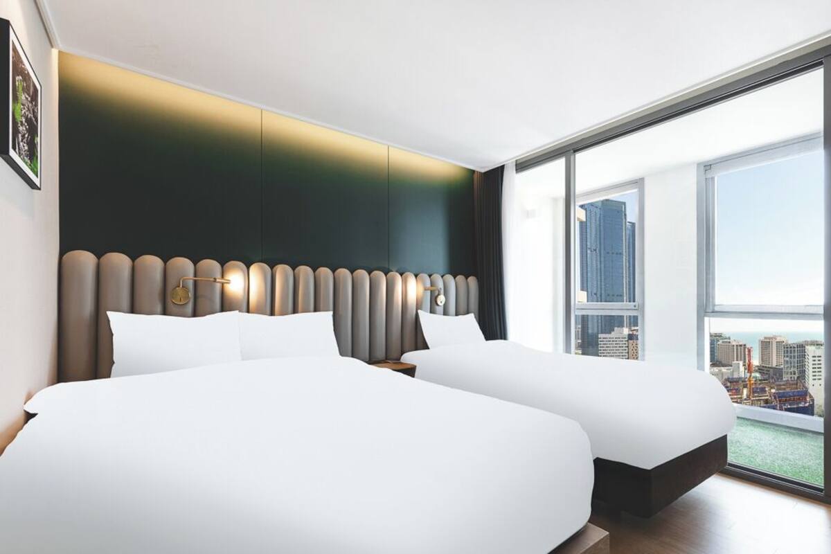Ein modernes Hotelzimmer mit zwei Betten, einem großen Fenster mit Stadtblick und einem außergewöhnlichen Kopfteildesign.