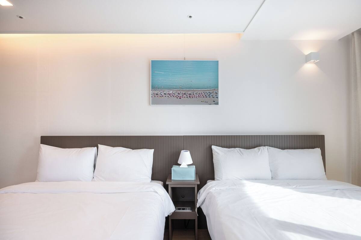 Ein Hotelzimmer mit zwei Betten, einem Nachttisch mit Lampe, einem Strandbild und einer Deckenleuchte.