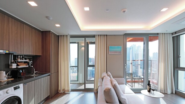 A modern hotel room with a kitchen, a sofa, and a balcony with a city view.