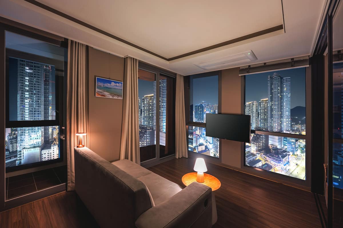 Ein modernes Hotelzimmer mit großem Fenster und Blick auf die Stadt, einer bequemen Couch, einem kleinen runden Tisch mit Lampe und einem flachbildfernseher an der Wand.