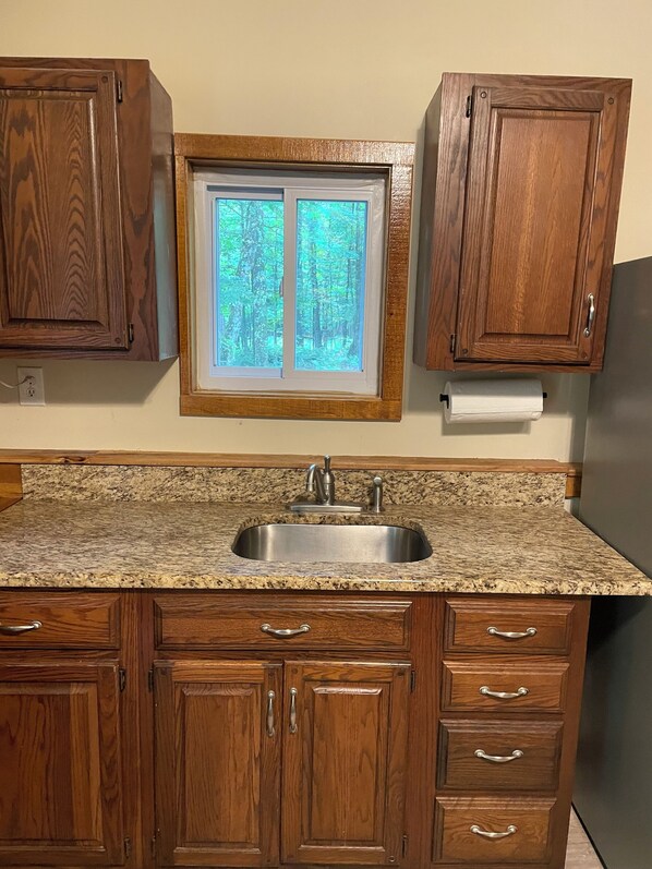 A kitchen with wooden cabinets, a granite countertop, a stainless steel sink, and a window with a view of trees.