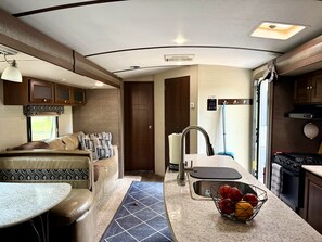A compact RV interior with a kitchenette, a seating area with a table, and a window providing natural light.