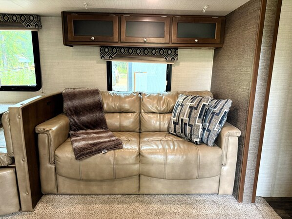 A tan leather sofa with a brown throw blanket and a patterned pillow, a window with a view of greenery, and a wooden cabinet above.