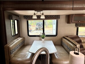 A cozy RV interior with a dining area, two cushioned seating areas, a small table, and a window with a view of a vehicle and trees.