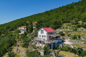 Propiedad en una ladera con varias construcciones, incluyendo una casa con techo rojo y un balcón, rodeada de vegetación y terreno rocoso.