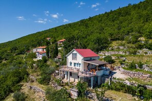 Una propiedad en una ladera con una casa grande con techo rojo, balcón y zona de piscina.