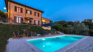 A two-story brick house with a swimming pool, outdoor seating, and a hillside backdrop.