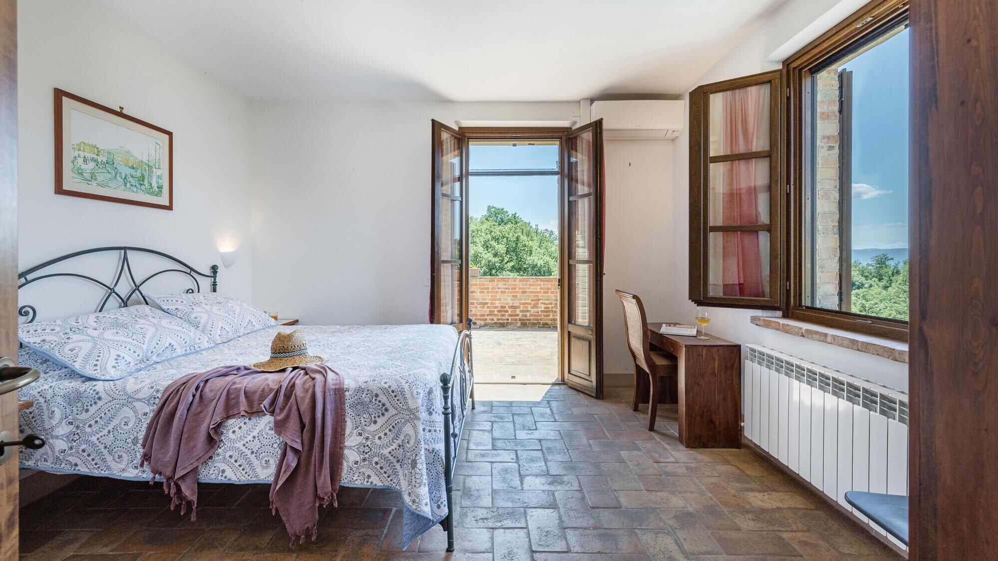 A bedroom with a bed, a chair, a table, and a window with a view of greenery.