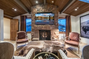 A stone fireplace with a TV above it, leather chairs, a fur rug, and a large window with a mountain view.
