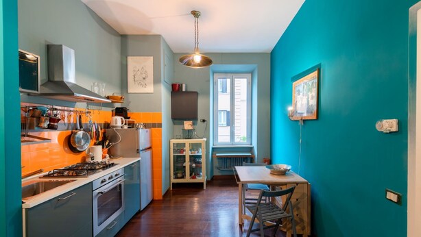 Een keuken met een gasfornuis, oven en kasten. Er is een raam met jaloezieën en een deur die naar een andere kamer leidt.