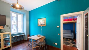 Een kamer met een blauwgroene wand, een houten tafel met een kom, een raam en een deur die leidt naar een blauwe kamer met een bank.