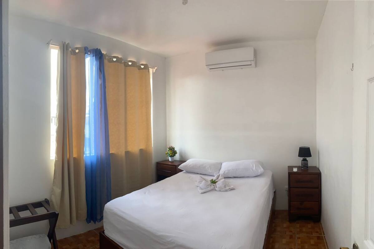 A hotel room with a bed, a nightstand, a lamp, and an air conditioning unit.
