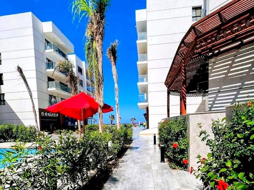 Un sendero bordeado por abundante vegetación y un paraguas rojo, que conduce a un edificio moderno con balcones.