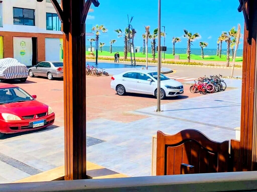 A parking area with cars and bicycles, a building with a modern facade, and a view of the beach with palm trees.