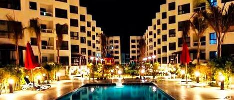 A swimming pool surrounded by multi-story buildings with balconies, outdoor seating, and palm trees.