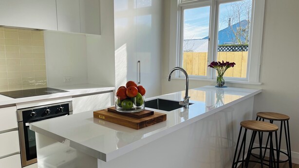 A modern kitchen with a white island, stainless steel appliances, and a window with a view of a wooden fence and mountains. - Luxurious House Next to St Clair Beach (Dunedin)