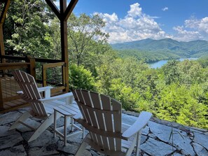 A covered outdoor seating area with Adirondack chairs overlooking a mountainous landscape and a lake.