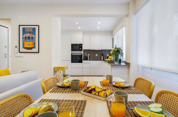 Cocina moderna con armarios blancos, un microondas y un lavamanos. Hay una mesa de comedor con alimentos para el desayuno y una silla amarilla.