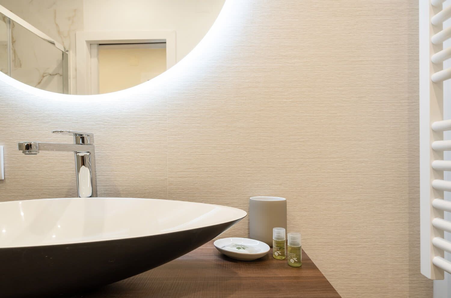 A bathroom with a large vessel sink, a mirror, a towel dispenser, and a wooden vanity with a plate and two bottles of lotion.
