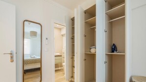 A hotel room with a wooden floor, a bed with a patterned bedspread, a large mirror, and a wardrobe with open doors revealing shelves and a hanging rack. - Monte Estoril Seabreeze by Portugal Portfolio (Estoril)