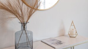 Un vaso di vetro trasparente con piante essiccate su un tavolo bianco, uno specchio rotondo con cornice dorata e un portacandele geometrico.
