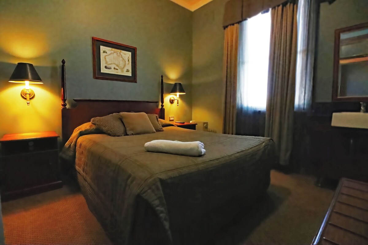 A neatly made bed with a white towel, two bedside lamps, a framed map on the wall, and a window with curtains.