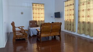 Una habitación con piso de madera, una mesa de comedor con sillas, un televisor en la pared y dos ventanas con cortinas estampadas.