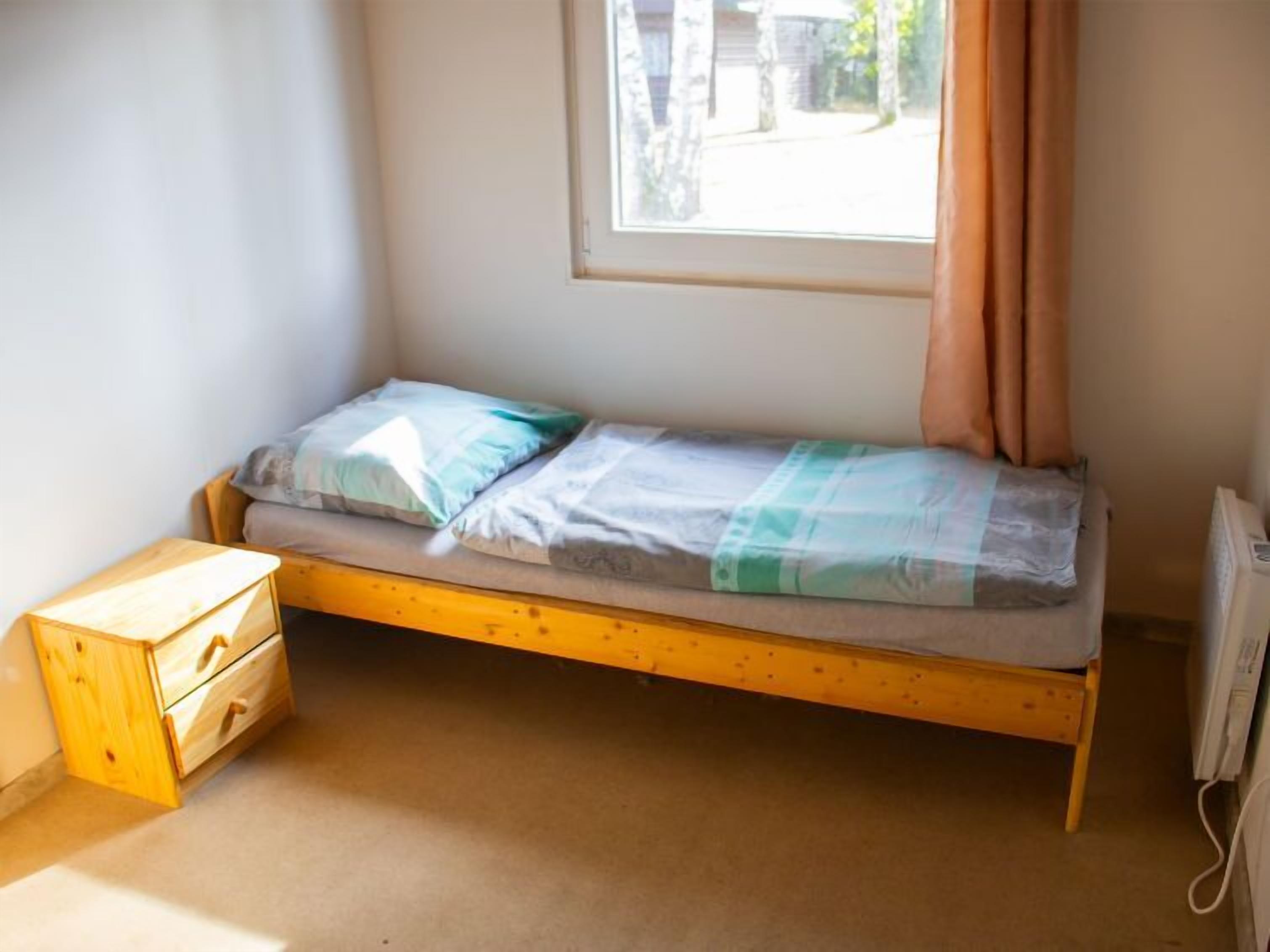 A small, single-bed room with a wooden frame, a wooden nightstand with drawers, and a window with curtains.
