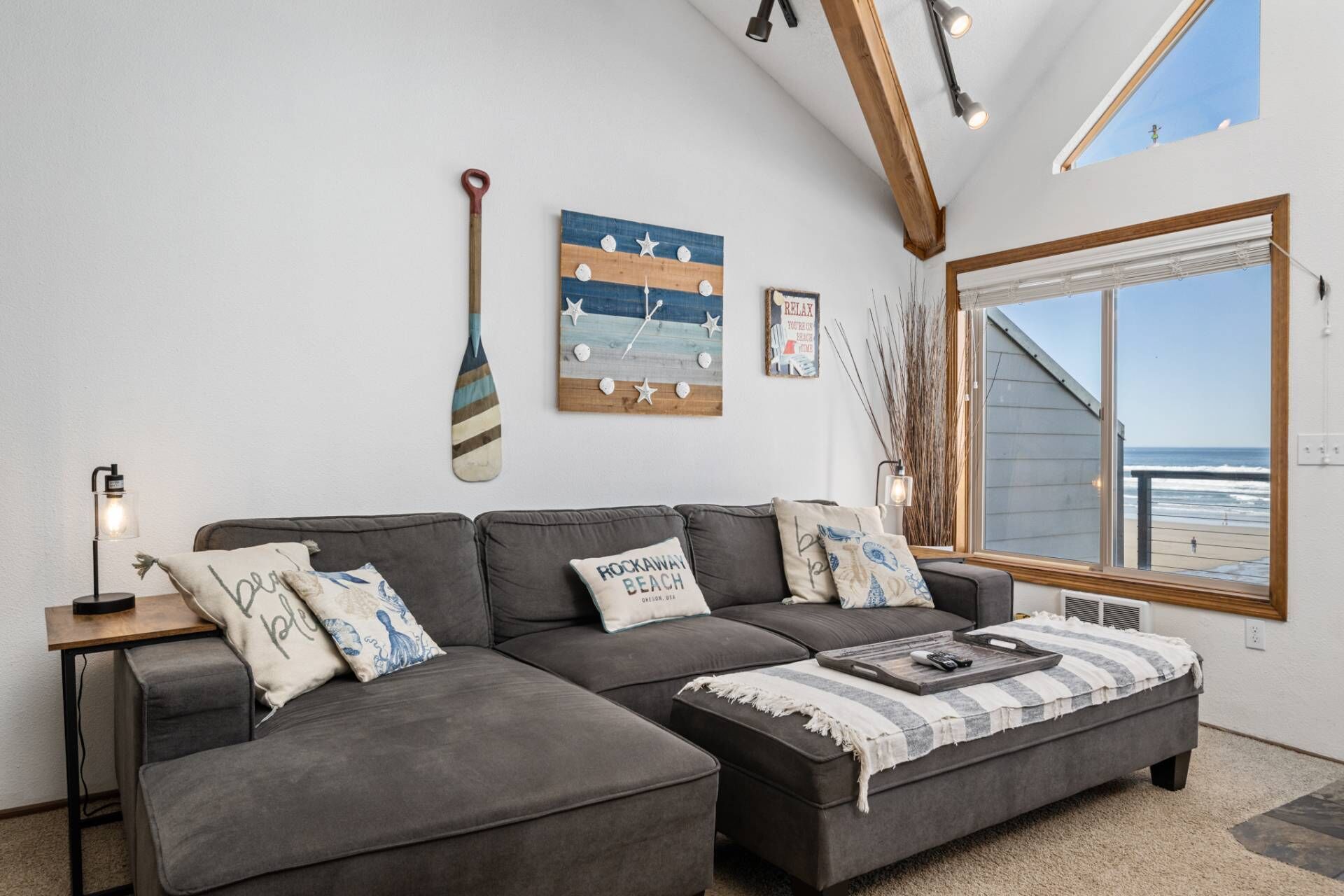 Ein modernes Wohnzimmer mit einer Eckcouch, Holzboden, einem großen Fenster mit Blick auf den Strand und maritimer Dekoration.