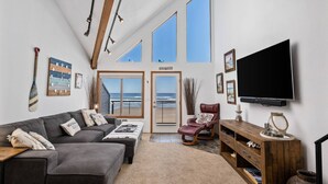 A spacious living room with a large gray sectional sofa, a leather recliner, a wooden entertainment unit, and a large TV. The room features a high ceiling with exposed beams, large windows offering a view of the ocean, and a door leading to a balcony. - Extraordinary Oceanfront Views at Rockaway Creek Inn, Patio and Living Area Bathed In Natural Light (Rockaway Beach)