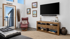 A living room with a large TV, a red chair, a wooden cabinet, and a view of the ocean through a glass door. - Extraordinary Oceanfront Views at Rockaway Creek Inn, Patio and Living Area Bathed In Natural Light (Rockaway Beach)