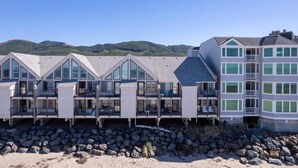A coastal hotel with multiple balconies, large windows, and a view of the beach and hills. - Extraordinary Oceanfront Views at Rockaway Creek Inn, Patio and Living Area Bathed In Natural Light (Rockaway Beach)
