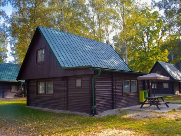 En stuga med ett sluttande tak av metall, ett picknickbord och en liten uteplats med gräs och träd i bakgrunden.